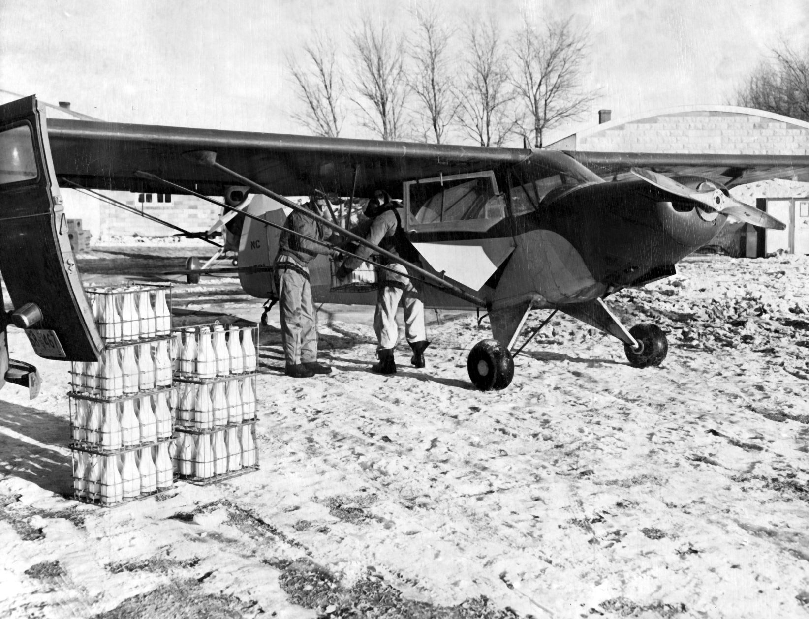 Dean Davis (left) and Red Meiners load milk into a plane at Ogallala for delivery to Grant, which had no dairy. Both men have flown many aid missions to ranches and isolated farms. Miners, manager of the Ogallala airport hauled in the body of an elderly man who died a natural death in Arthur County. Published Jan. 10, 1949.